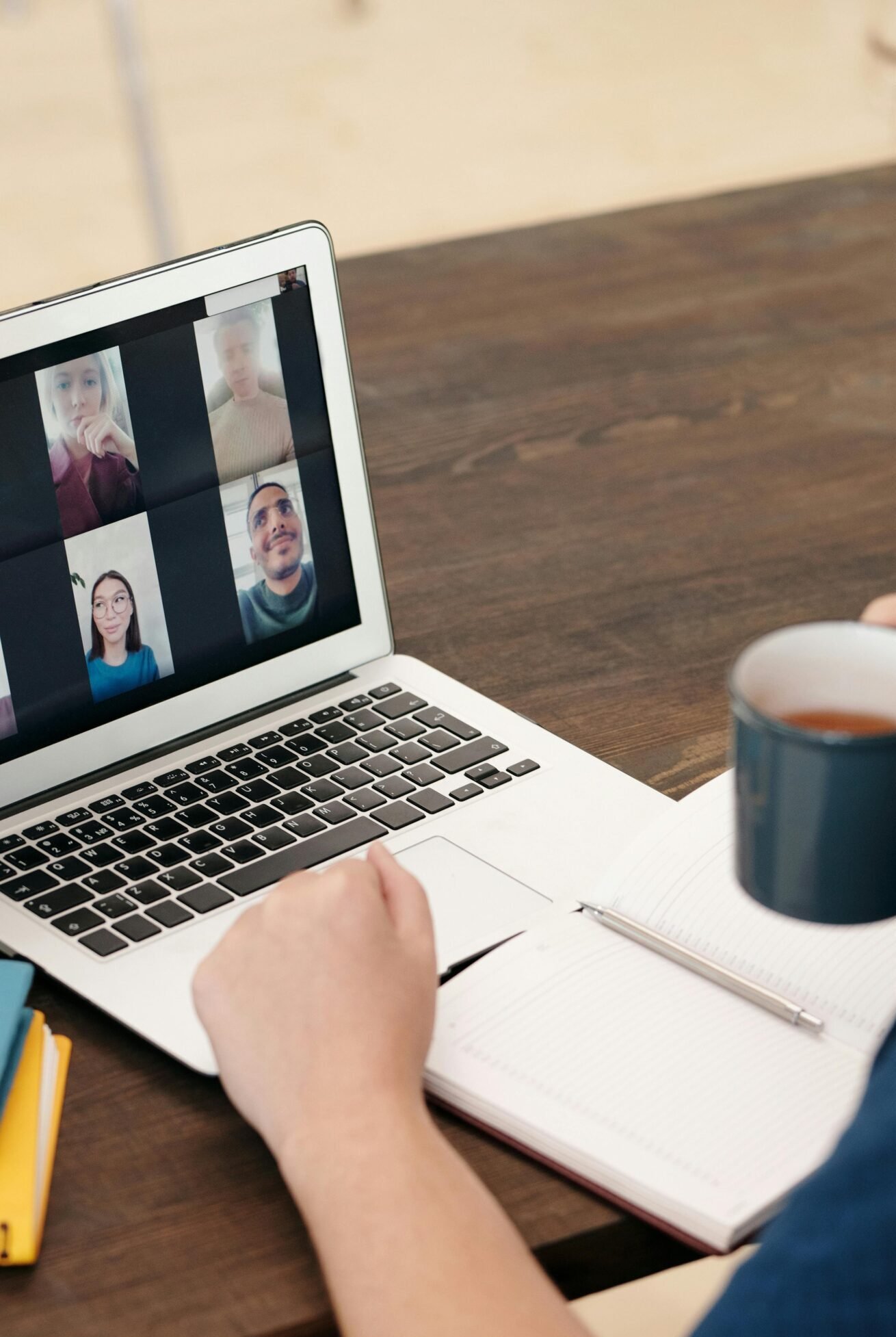 A person participates in a video conference call while drinking coffee at a desk with a laptop and notebook.
